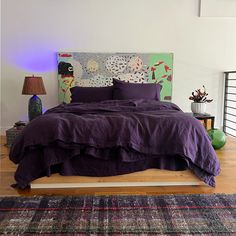 a bed with purple sheets and pillows in a room next to a painting on the wall