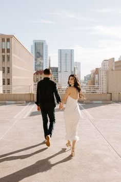 a man and woman walking in the city holding hands, with buildings in the background