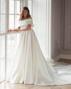 a woman standing in front of a window wearing a white wedding dress with off the shoulder sleeves