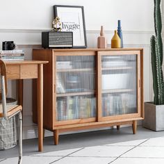 a wooden cabinet with glass doors next to a desk and cactus in a pot on the floor