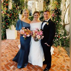 three people standing next to each other on a brick floor with flowers in the background