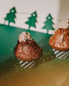 two chocolate muffins sitting next to each other on a green surface with christmas trees in the background