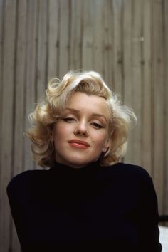 a woman with blonde hair sitting in front of a wooden wall wearing a black top