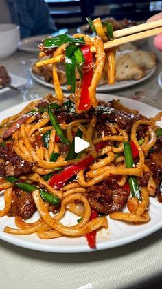 someone holding chopsticks over a plate of noodles with meat and vegetables on it