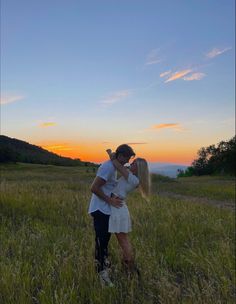 a man and woman standing in the middle of a field at sunset with their arms around each other