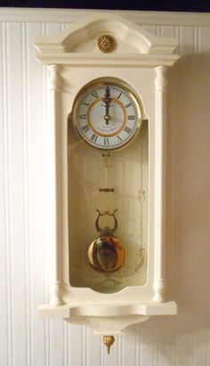 a white clock mounted to the side of a wall next to a bell and pendulum