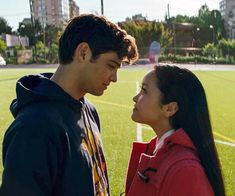 a young man and woman standing in front of a soccer field looking at each other