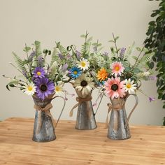 three metal vases with flowers in them on a wooden table next to a potted plant