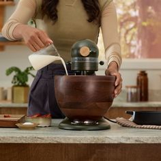 a woman pouring milk into a wooden bowl on top of a kitchen counter next to an electric mixer