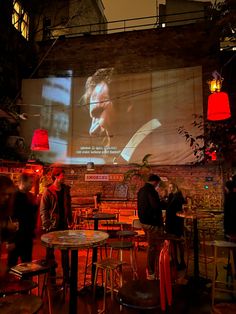 people are standing in front of a large screen at an outdoor restaurant with red lights