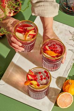 two people holding glasses filled with fruit on top of a green tablecloth next to sliced oranges