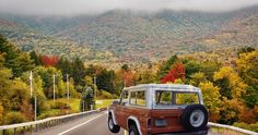 an old jeep driving down the road in front of a mountain range with fall foliage