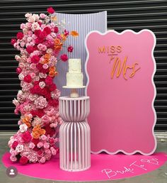 a cake on a stand next to a pink sign and flower arrangement in front of it