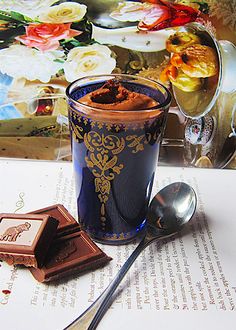 a cup of chocolate next to a spoon on top of an open book with flowers in the background