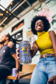 a woman holding up a can of beer in front of a man sitting on a bench