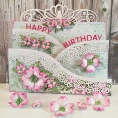 a happy birthday card with pink flowers and lace on the edge, next to some petals