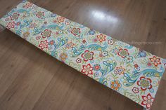 an upholstered bench cushion with colorful flowers and peacocks on white fabric sitting on a wooden floor