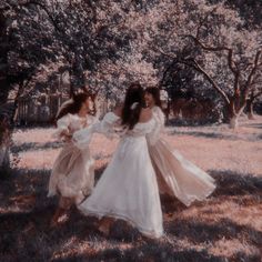 two women in white dresses are walking through the grass together, with trees behind them