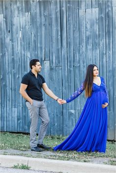 a pregnant woman in a blue dress holds the hand of her husband as they stand next to a wooden fence