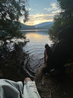 someone's feet in the water with trees and mountains in the background at sunset