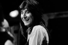 a black and white photo of a smiling man with long hair, wearing a collared shirt