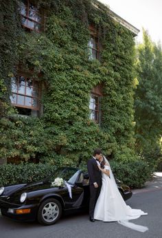 a bride and groom standing in front of a black sports car