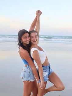 two young women standing on the beach hugging each other and smiling at the camera with their arms in the air