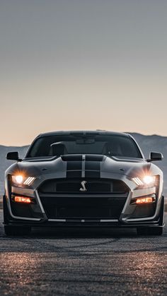 the front end of a black and white sports car with its lights on at dusk