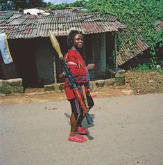 a woman in red jacket holding two large wooden sticks and walking down street with house behind her