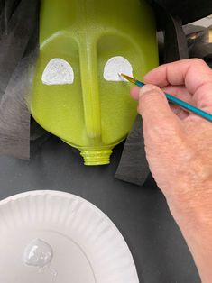 a person is painting a green face on a plastic container with a pencil and paper plate next to it