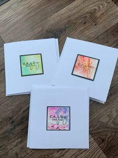 three cards with watercolor designs on them sitting on a wooden floor next to a pair of scissors