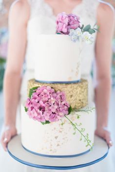 a woman holding a three tiered cake with pink flowers on the top and bottom