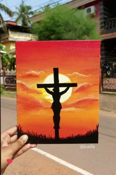 a hand holding up a painting of a person on the cross in front of a sunset