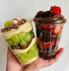 two desserts in plastic containers with fruit and chocolate toppings on top, one being held by a person's hand