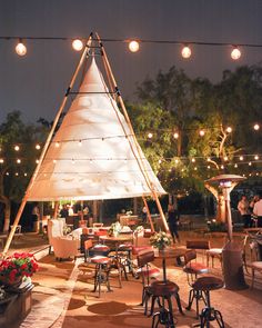 an outdoor dining area is lit up with string lights and wooden tables, chairs, and stools