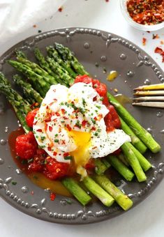 asparagus with tomatoes and poached eggs on a gray plate next to some dipping sauce