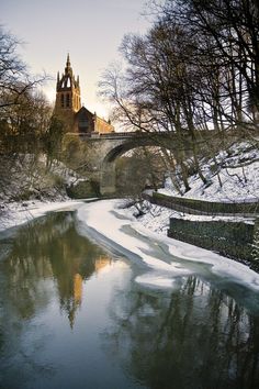 a river running through a snow covered forest next to a bridge with a church in the background