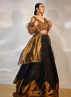 This exquisite Black and Gold Embellished Lehenga Set is a stunning fusion of traditional elegance and contemporary style, meticulously crafted in luxurious tissue and taffeta silk. The black lehenga, adorned with intricate hand embellishments, exudes opulence. Paired with the blouse features an alluring neckline and detailed craftsmanship. The look is completed by a gracefully draped dupatta, adding a final touch of sophistication. Ideal for grand celebrations such as Cocktails or Sangeet ceremonies, this ensemble radiates charm and grace. Pair it with statement jewellery and heels to elevate your look, ensuring you stand out on any occasion. Composition : Blouse, Lehenga and Dupatta - Taffeta Silk Care: Dry Clean Only and Vacuum Storage This product can be customized for sleeves, blouse