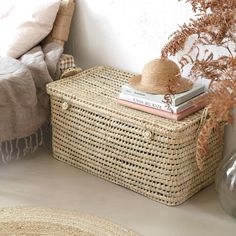 a wicker basket with books on it next to a bed and plant in a vase