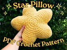 a hand holding a yellow crochet star pillow in front of a tree with stars on it