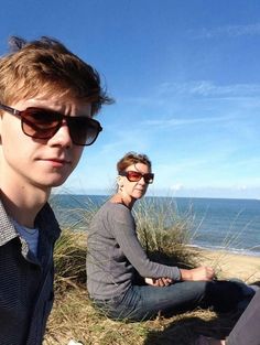 two people are sitting on the sand near the ocean and one person is taking a selfie