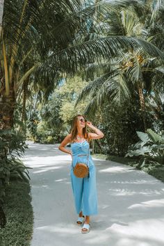 a woman standing in front of palm trees wearing a blue jumpsuit and straw bag