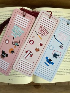 three bookmarks with stickers on them are sitting next to an open book and tassel