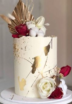 a white cake with gold leaf and flowers on top is sitting on a plate in front of a window