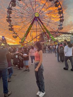 a woman standing in front of a ferris wheel at an amusement park as the sun sets