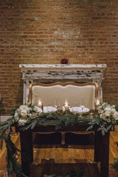 a table with candles and flowers on it in front of a brick wall, decorated with greenery