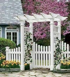 a white garden arbor with flowers in the foreground and a house in the background