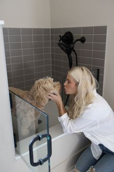 a woman petting a dog in a bath room next to a shower head and hand rail