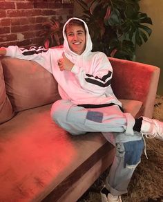 a man sitting on top of a couch wearing a pink and black hoodie with his thumb up