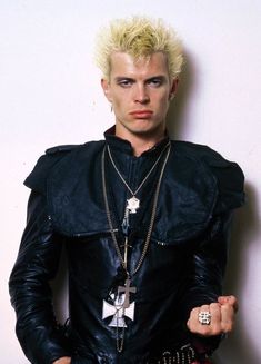 a man with blonde hair wearing a black leather jacket and chain necklace, standing in front of a white wall